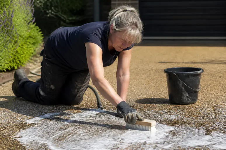 Femme nettoyant une allée de garage en résine avec une brosse et de l'eau savonneuse, entretien facile.