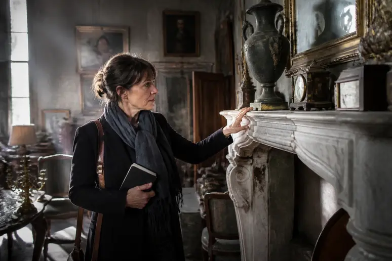 Une femme d'âge mûr observe attentivement un manteau de cheminée en marbre ancien dans une boutique d'antiquités parisienne.