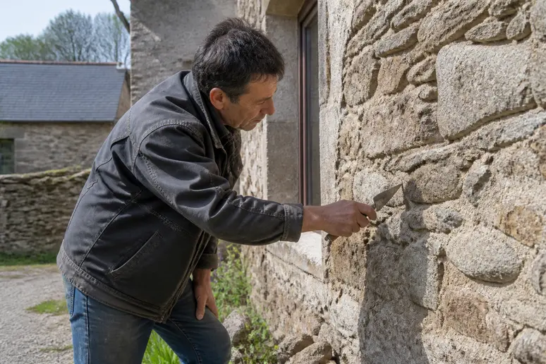 Un maçon français examine attentivement la façade en pierre d'une maison rurale traditionnelle sous le soleil.