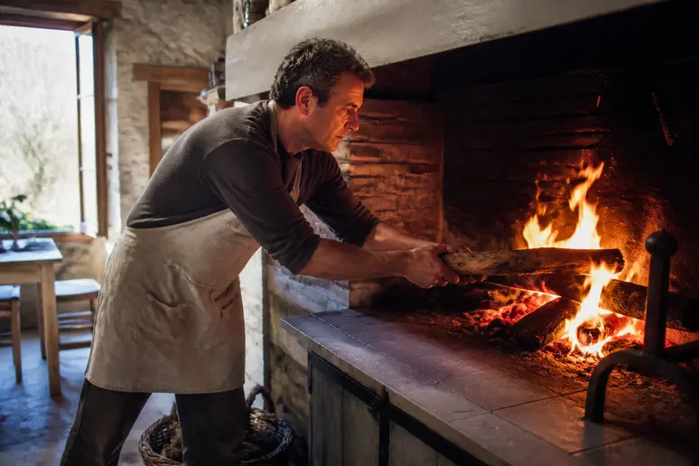 Un homme d'âge mûr charge du bois dans un imposant poêle de masse dans une cuisine de ferme ensoleillée.