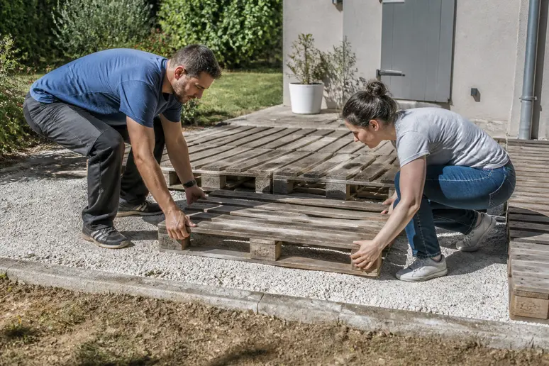 Deux personnes assemblent des palettes Europe (EUR/EPAL) pour former la structure d'une terrasse sur un lit de gravier.